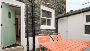 A patio with a table and chairs, a stone wall, a green door, and a white window. - Capella Cottage (Keswick)