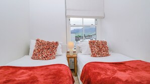 A room with two beds, each with a red blanket and a floral pillow, a window with a view of the outside, and a white wall. - Capella Cottage (Keswick)