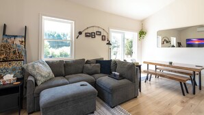 Een moderne woonkamer met een hoekbank, een eettafel met banken, een groot raam en een aan de muur bevestigde televisie.