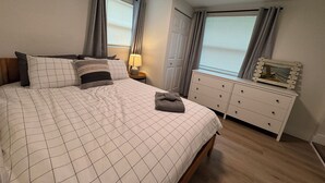 A bedroom with a bed, bedside table, lamp, dresser, and windows with curtains.