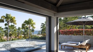 A modern room with a large window overlooking a rocky beach and palm trees. There is a lounge chair with a cushion and a table under a large umbrella. - Lagos del Mar (Punta de Mita)