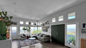 A spacious living room with a large gray sectional sofa, a coffee table, and a potted plant. There are multiple windows providing natural light and a view of the cityscape. - Mountain Top 4 Bdrm Home Along Nature Reserve with Spectacular Views, AC & WiFi (Spring Valley)