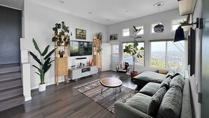 A modern living room with a grey sofa, a flat-screen TV, and a large window offering a city view. - Mountain Top 4 Bdrm Home Along Nature Reserve with Spectacular Views, AC & WiFi (Spring Valley)
