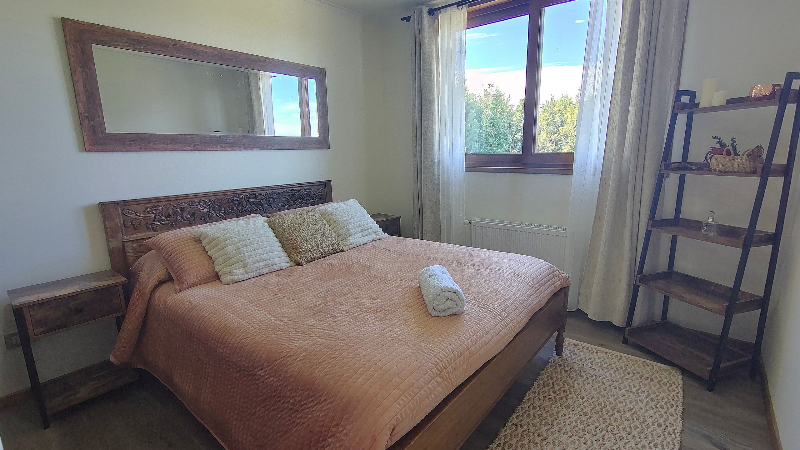 Une chambre à coucher avec un lit, une table de chevet, un miroir, une fenêtre avec des rideaux et une étagère en bois.