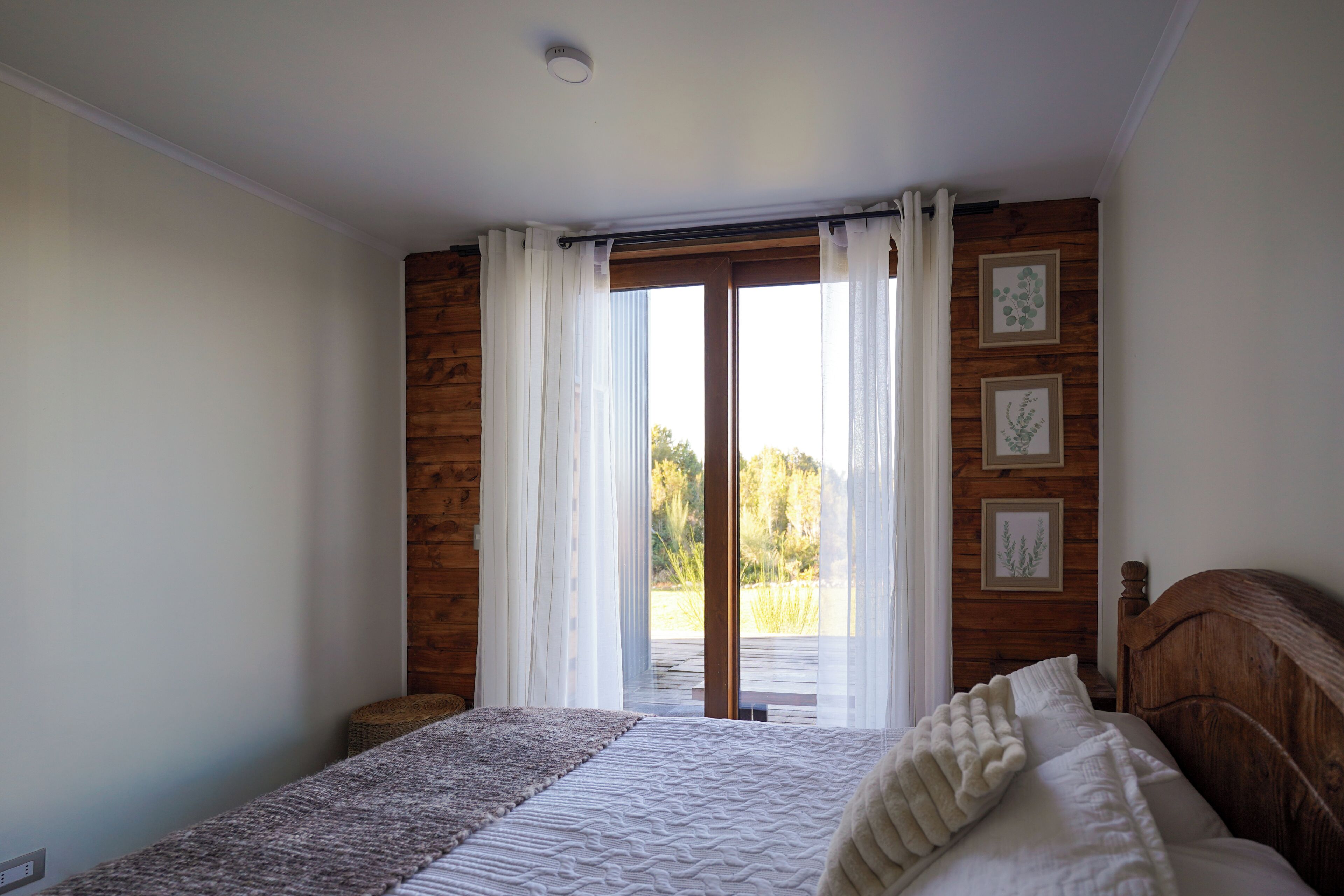 Een slaapkamer met een houten hoofdeinde, een bed met wit beddengoed, een raam met gordijnen en uitzicht op groen.