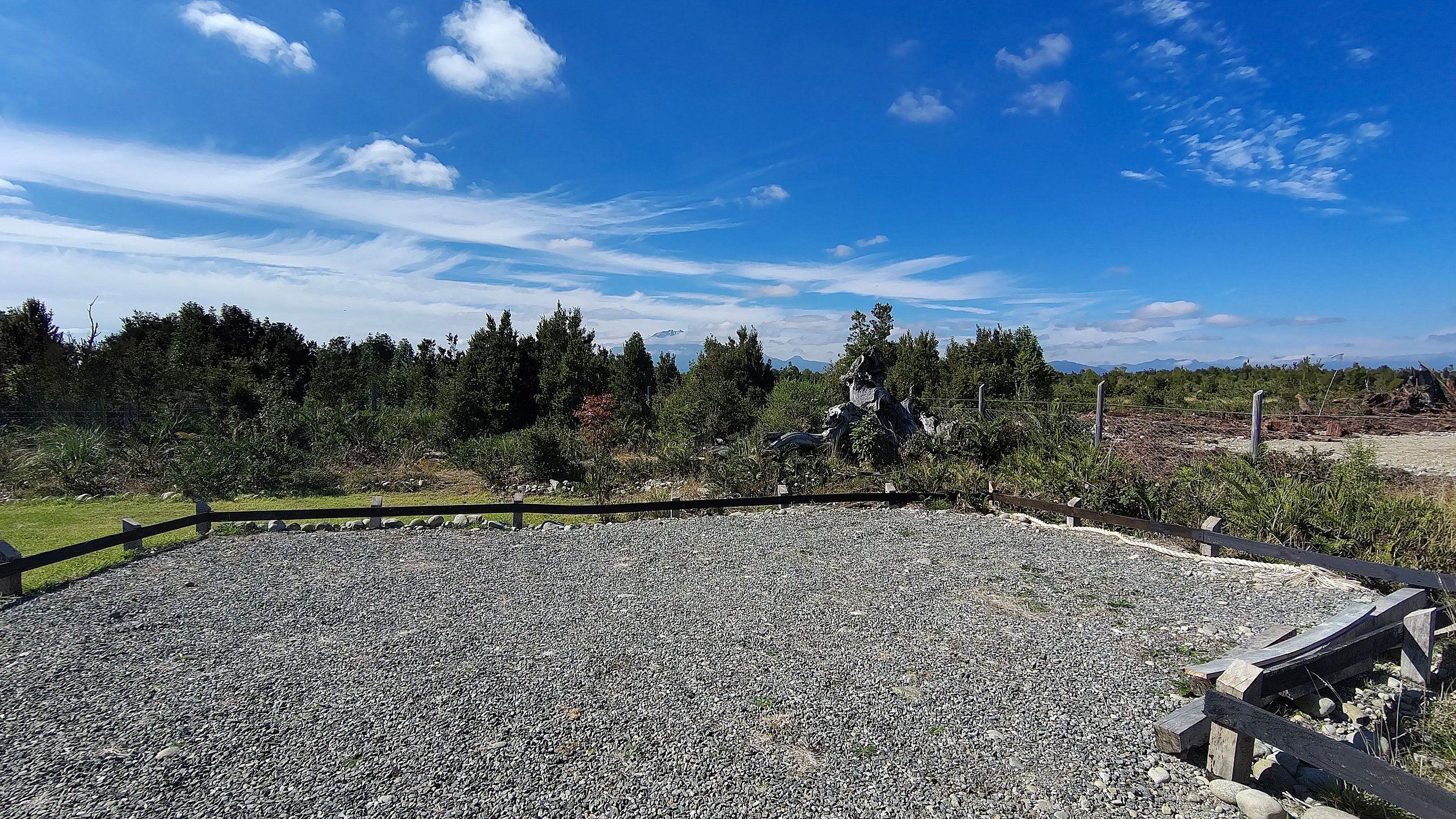 An outdoor area with a gravel surface, surrounded by a wooden fence, and a backdrop of trees and mountains under a clear sky.
