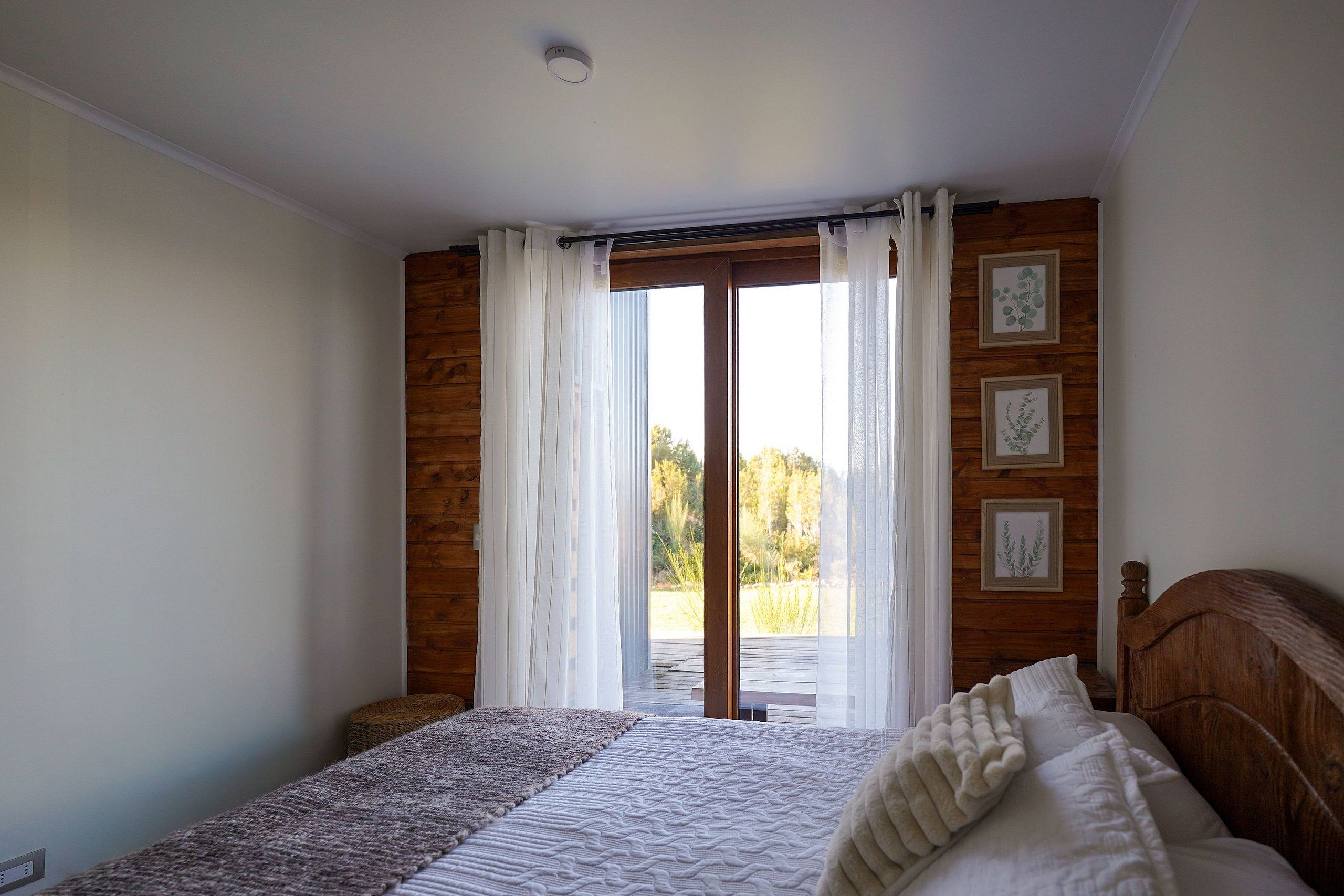 Un dormitorio con cama, cabecera de madera, ventana con cortinas y vistas al exterior.