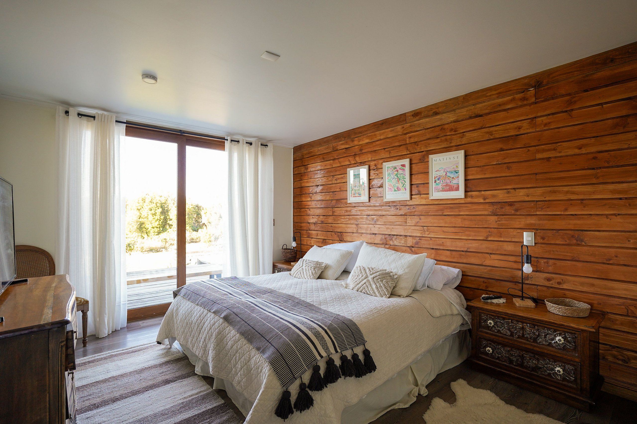 Un dormitorio con pared de madera, cama con ropa de cama blanca, mesita de noche, lámpara y ventana con cortinas.
