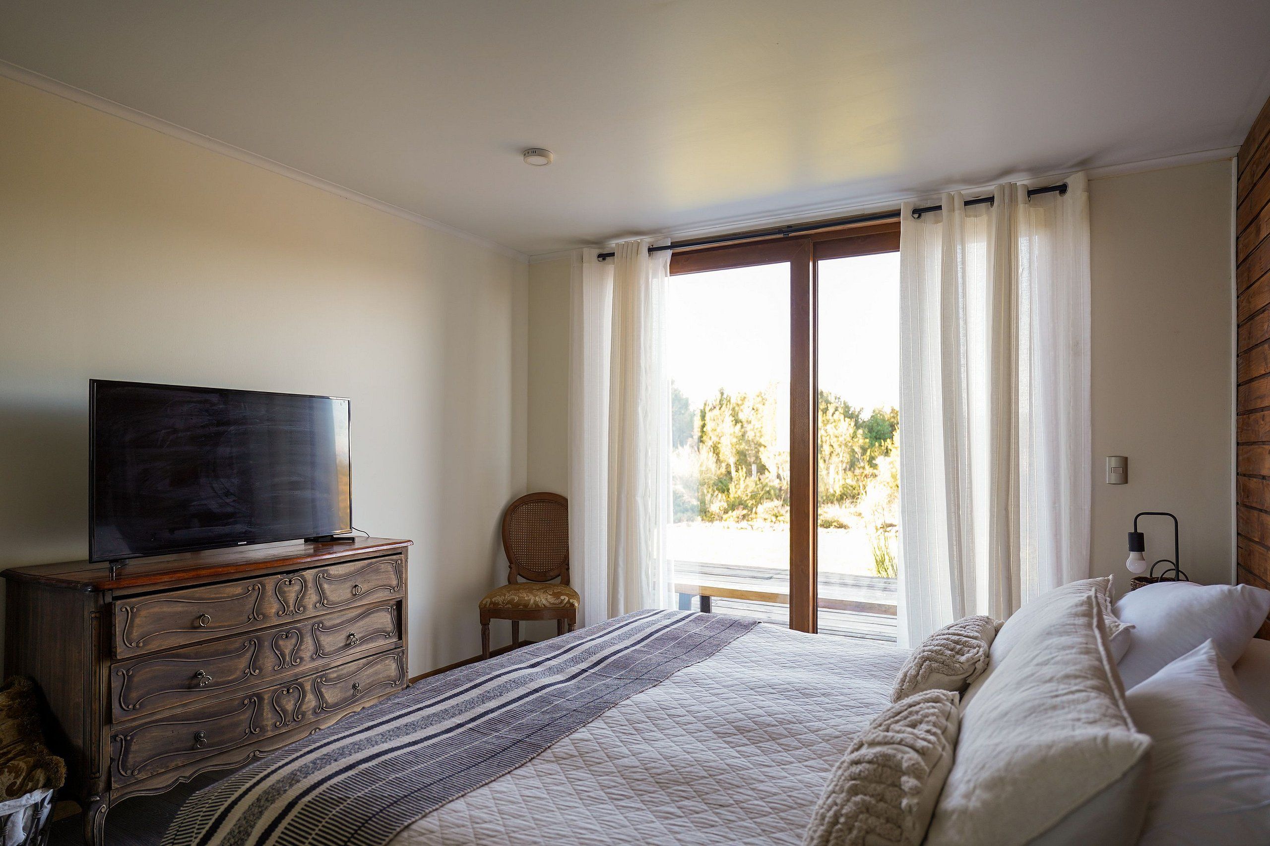 A bedroom with a bed, a television on a dresser, a chair, and a window with curtains.