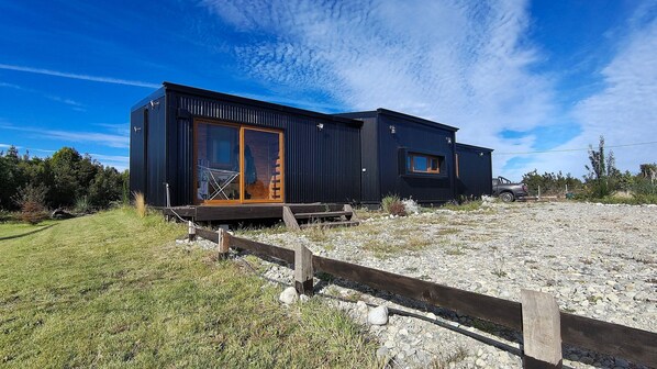 A modern, black, modular building with large windows and a wooden door, situated on a gravel area with a wooden fence in front.