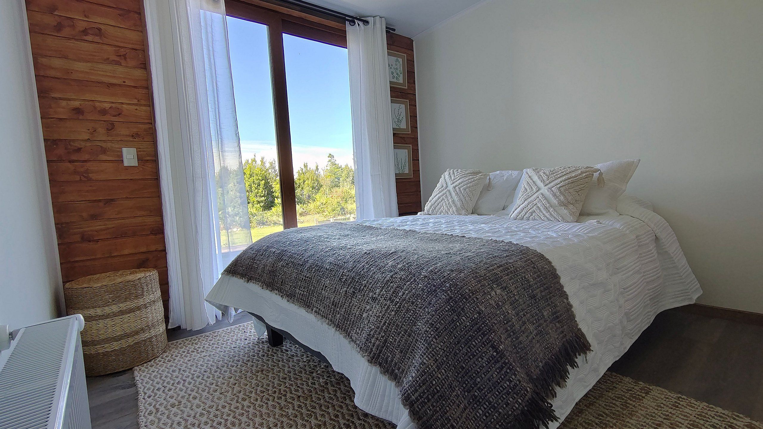 Quarto com cama, janela com cortinas, radiador e cesta de palha.