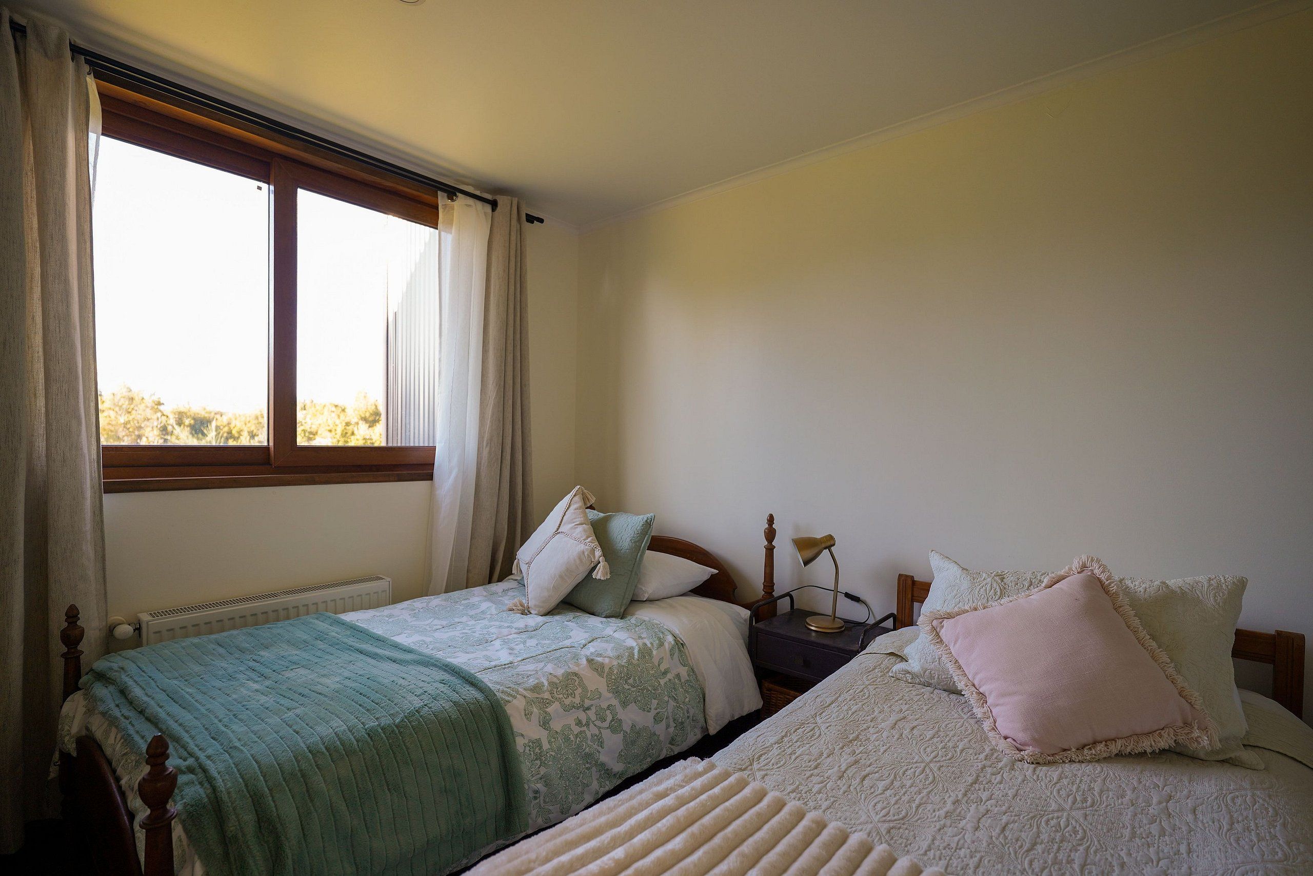 A bedroom with two beds, a window with curtains, a radiator, and a bedside lamp.
