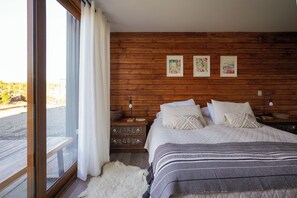 A bedroom with a wooden headboard, a large bed with white bedding, a nightstand with a lamp, and a view through a window with curtains.