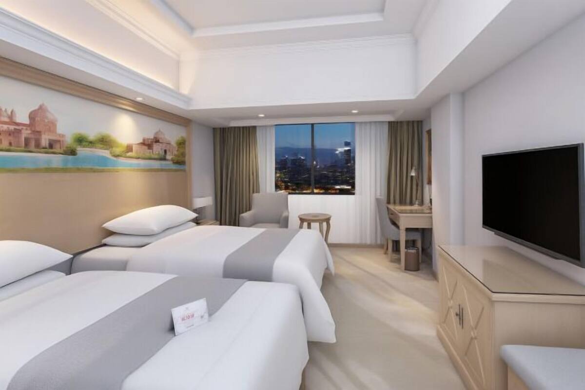 A hotel room with two beds, a desk, a TV, and a city view.