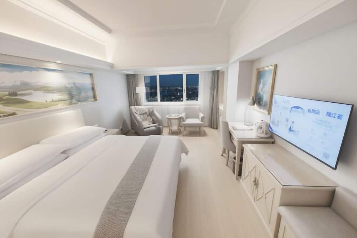 A hotel room with a large bed, a desk with a chair, a television, and a window with a city view.