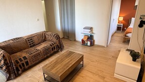 A living room with a sofa, a coffee table, a small side table with books, and a door leading to another room. - Perle de Carnon Plage (Mauguio)