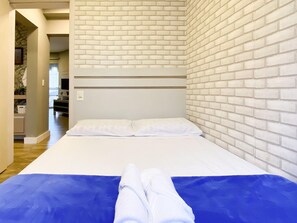 Una cama con ropa de cama blanca y un detalle azul, una pared de ladrillo y un pasillo que conduce a un baño.