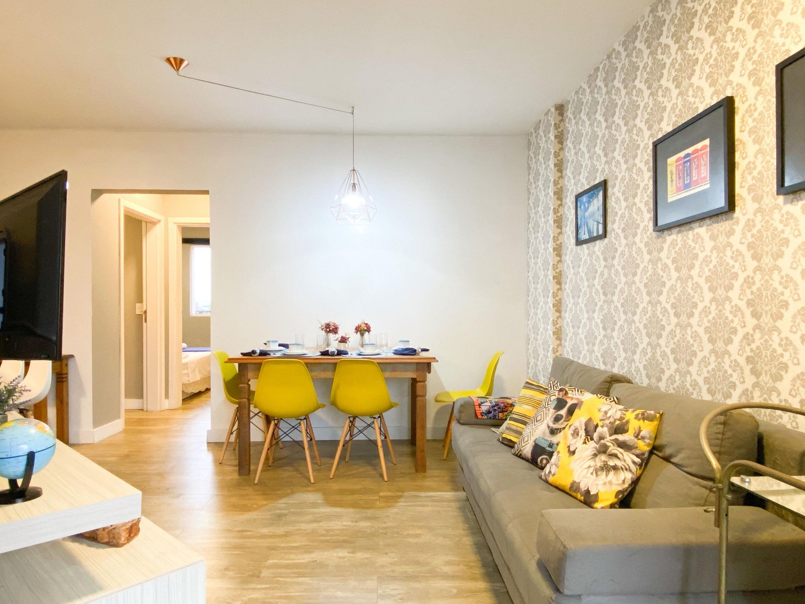 A modern living room with a grey sofa, yellow chairs, a wooden dining table set for four, and a TV mounted on the wall.