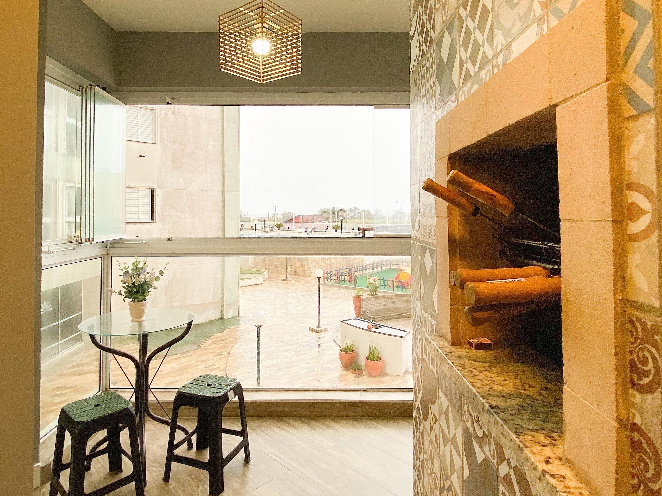 Uma varanda com mesa de vidro e dois banquinhos, lareira e vista para uma piscina e edifícios.