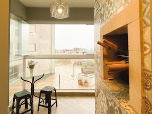 Un balcón con una mesa de vidrio y dos taburetes, una chimenea, y vistas a una piscina y edificios.