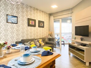 Een moderne woonkamer met een bank, een eettafel gedekt met borden en kopjes, een aan de muur bevestigde televisie, en een balkon met uitzicht.