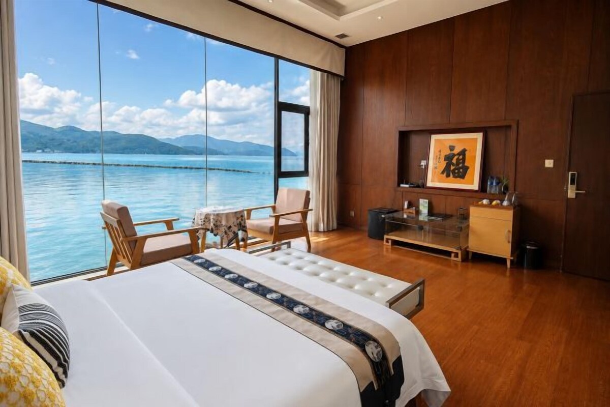 A hotel room with a large bed, a desk, a chair, a small table, and a view of the sea.