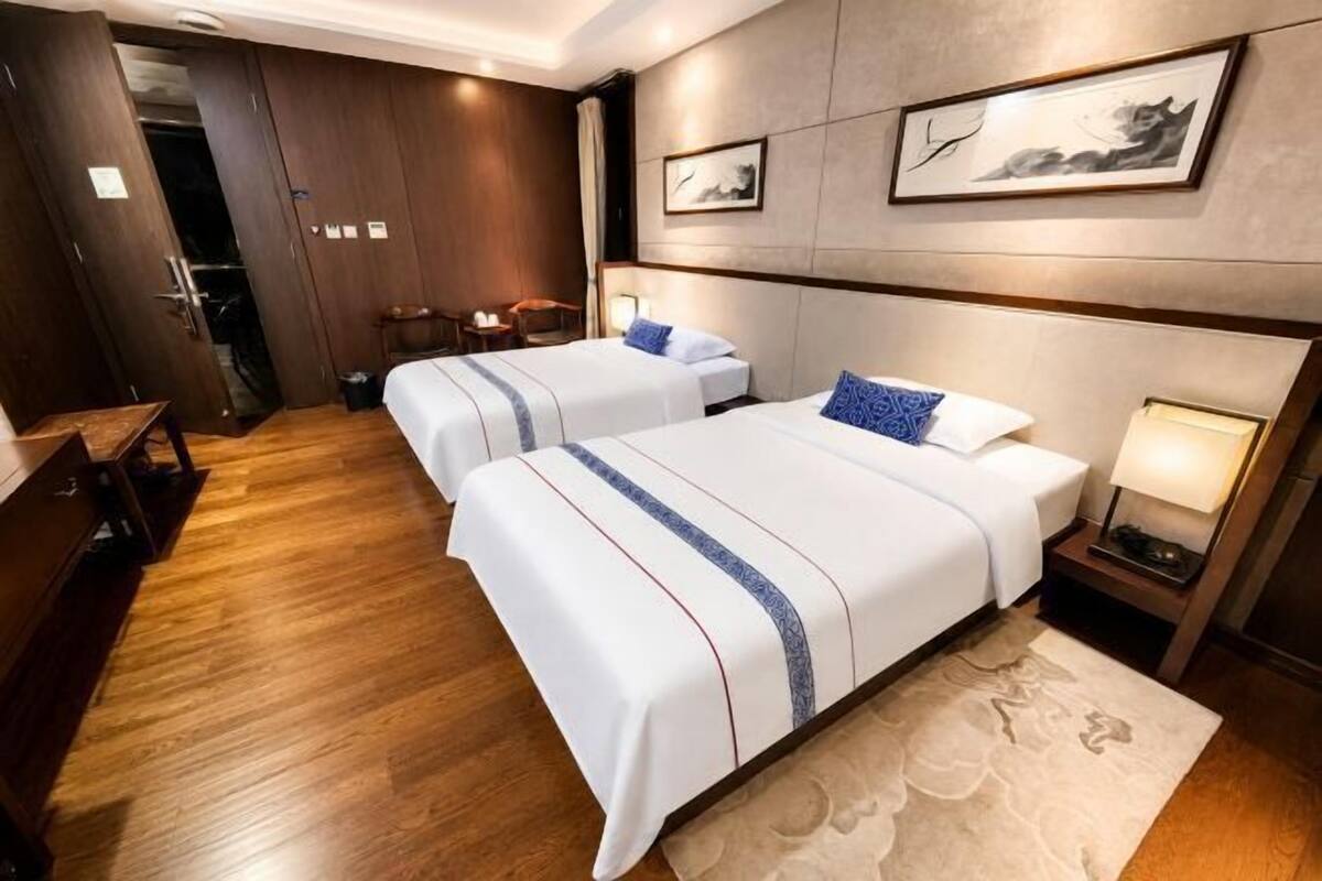A hotel room with two beds, a wooden floor, a desk, a chair, a nightstand with a lamp, and artwork on the wall.