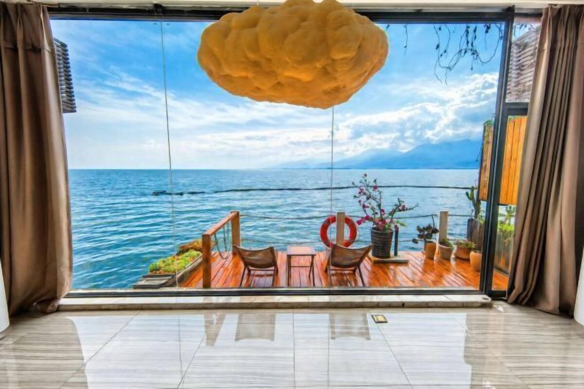 A room with a large window overlooking a body of water, a wooden deck outside, and a hanging light fixture.