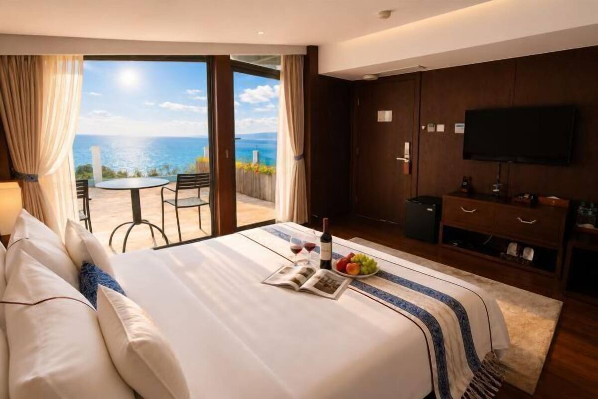 A hotel room with a bed, a dining table, a TV, and a view of the sea.