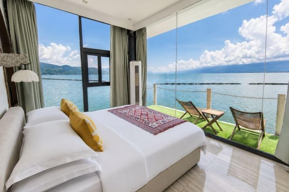 A hotel room with a large bed, a view of the sea, and a balcony with chairs.