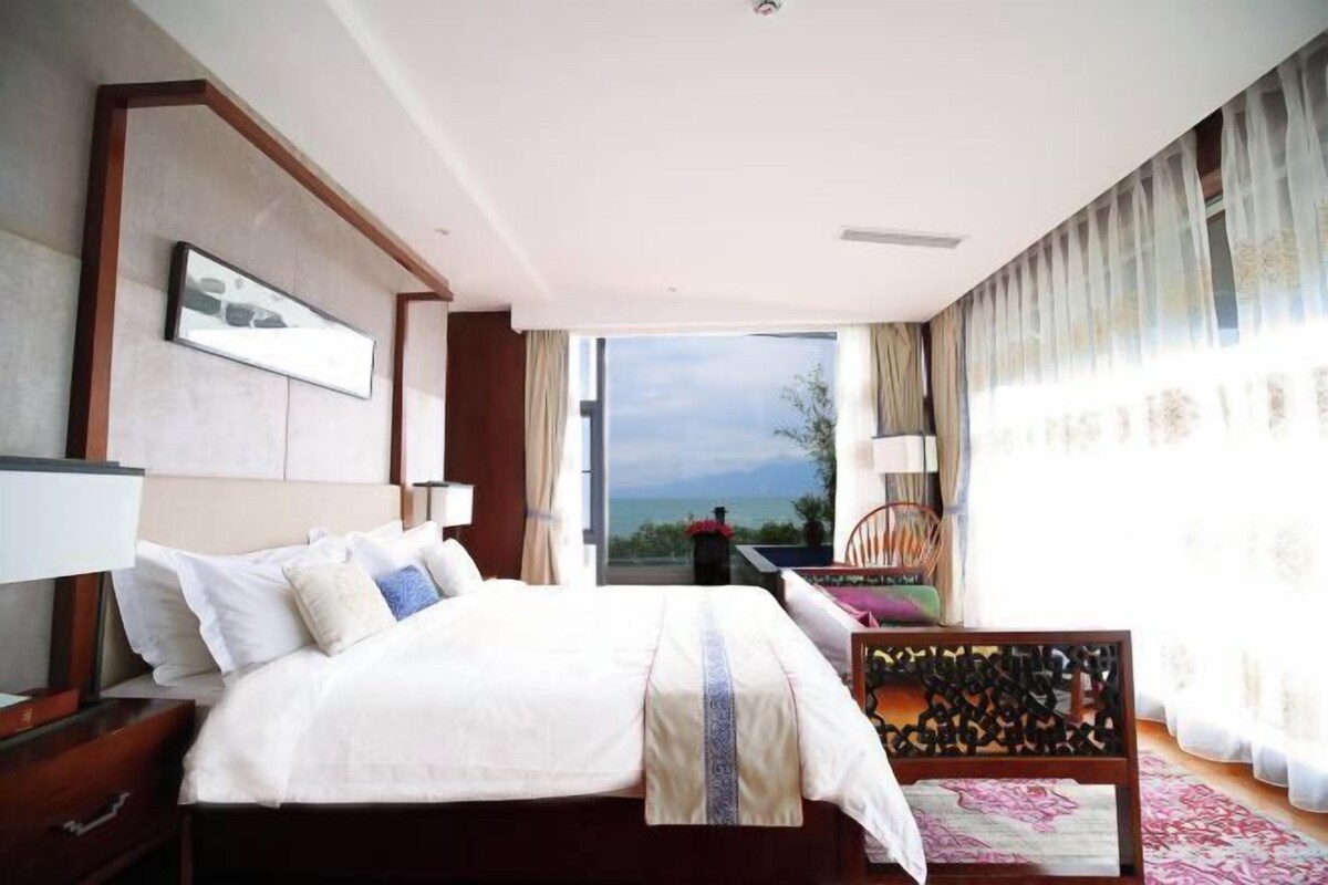 A hotel room with a large bed, a wooden headboard, a desk with a chair, a window with curtains, and a view of the outdoors.