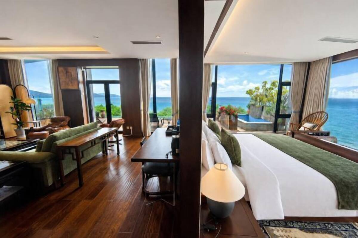 A modern hotel room with a large window, a sofa, a dining table, and a lamp.