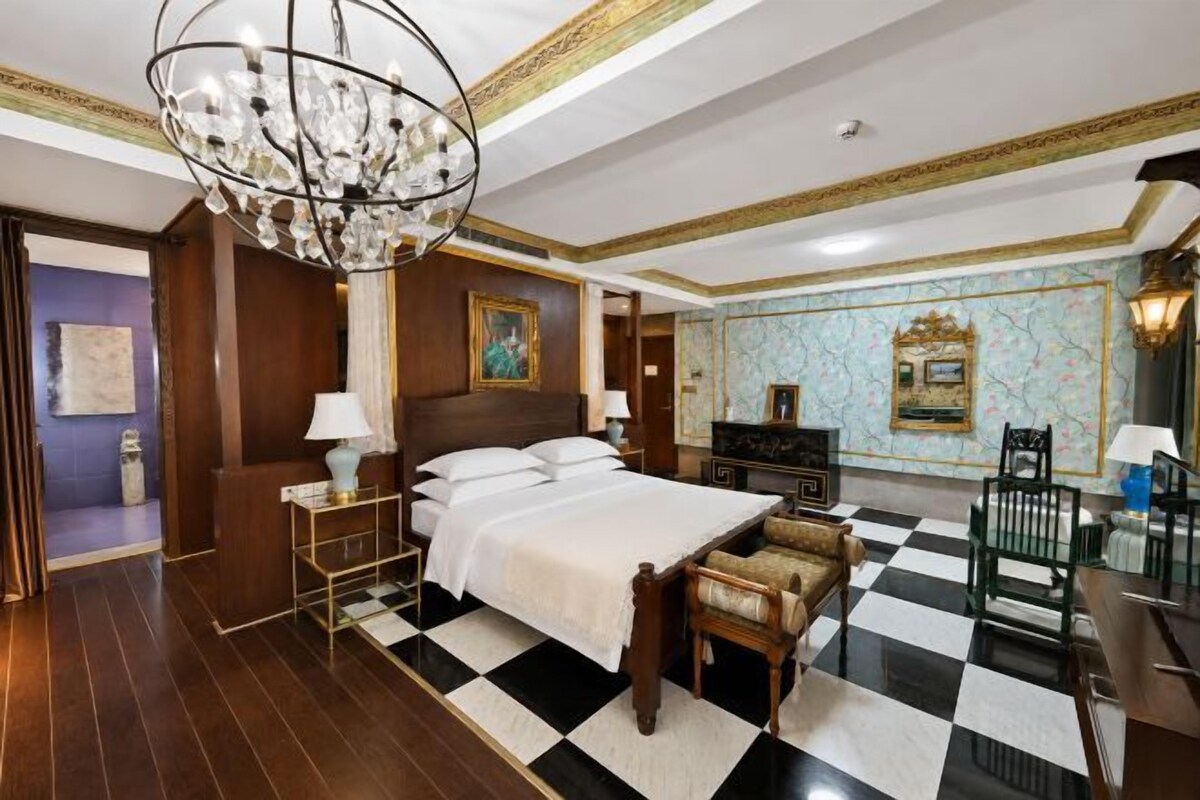 A hotel room with a large bed, a chandelier, a checkered floor, and a decorative wall.