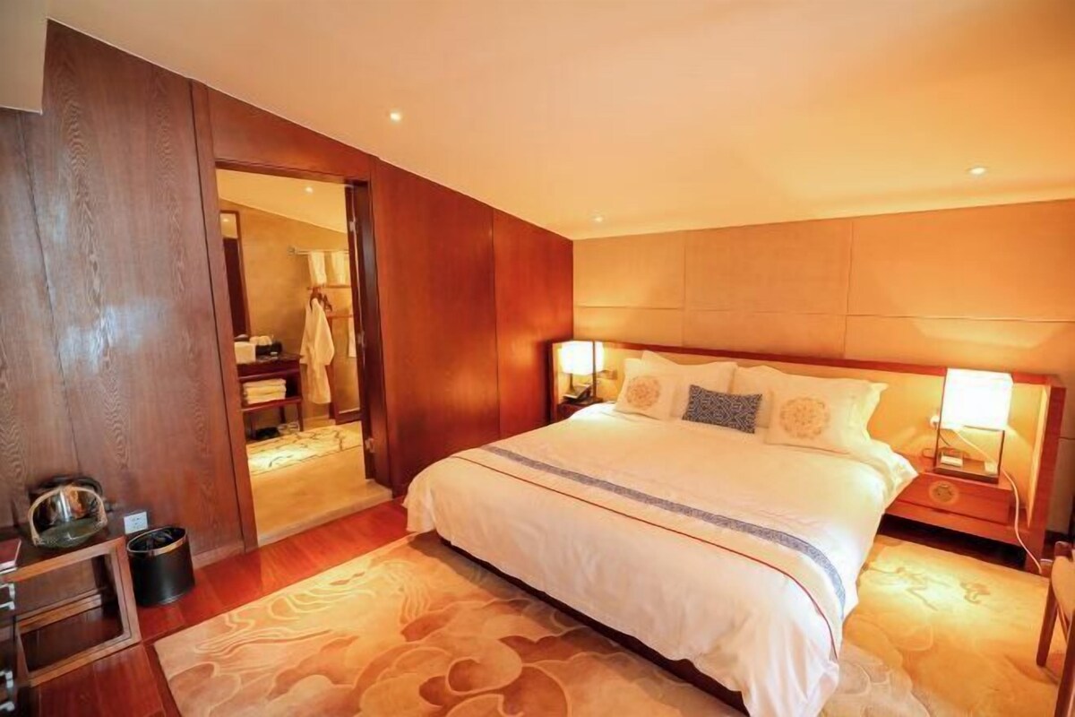 A spacious bedroom with a large bed, wooden paneling, a bedside table with a lamp, and a view into the bathroom.