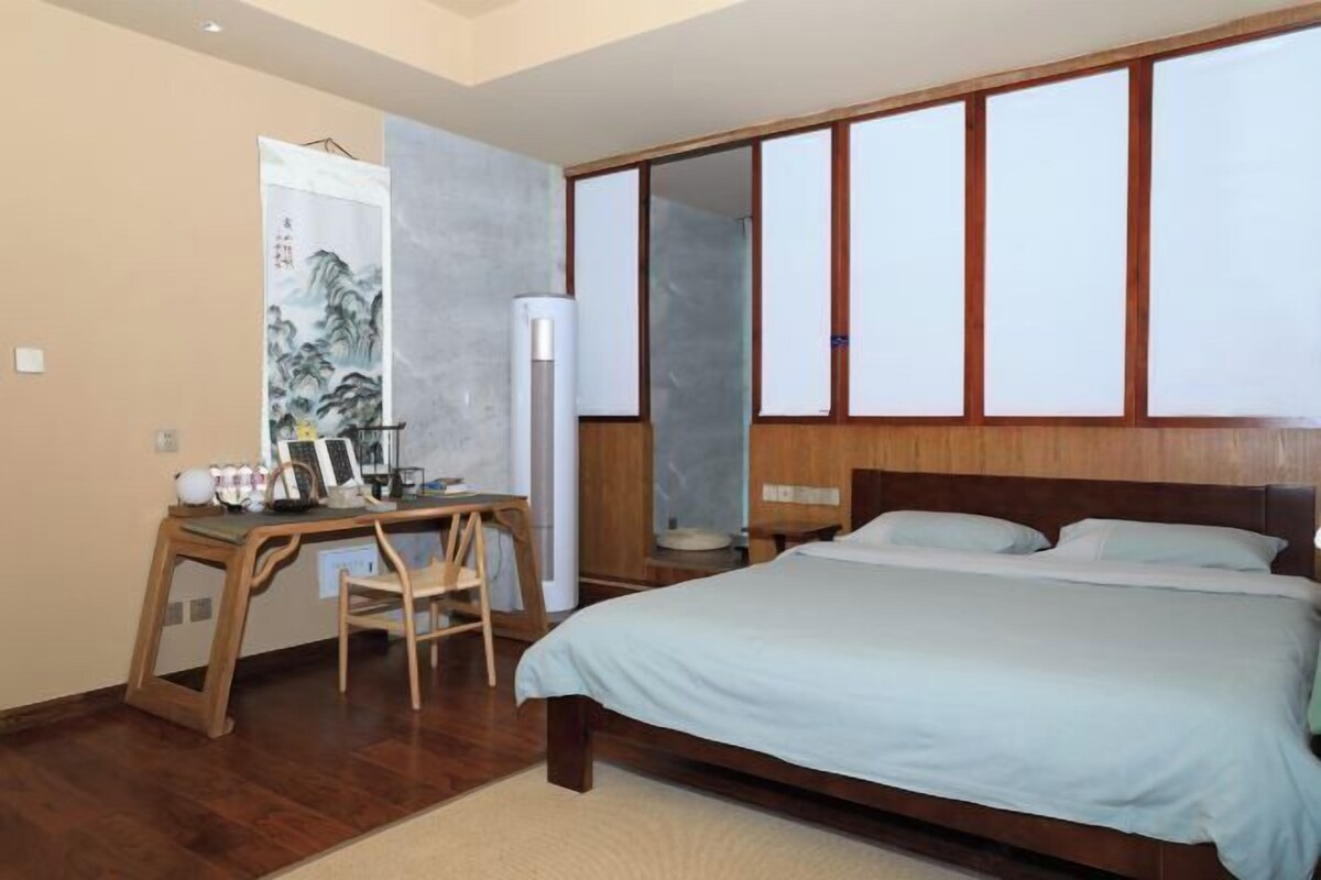 A bedroom with a bed, a desk with a chair, and a large window with wooden frames.