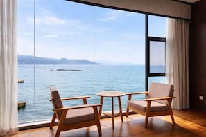 A room with a large window, two wooden chairs, a small round table, and a view of the sea.