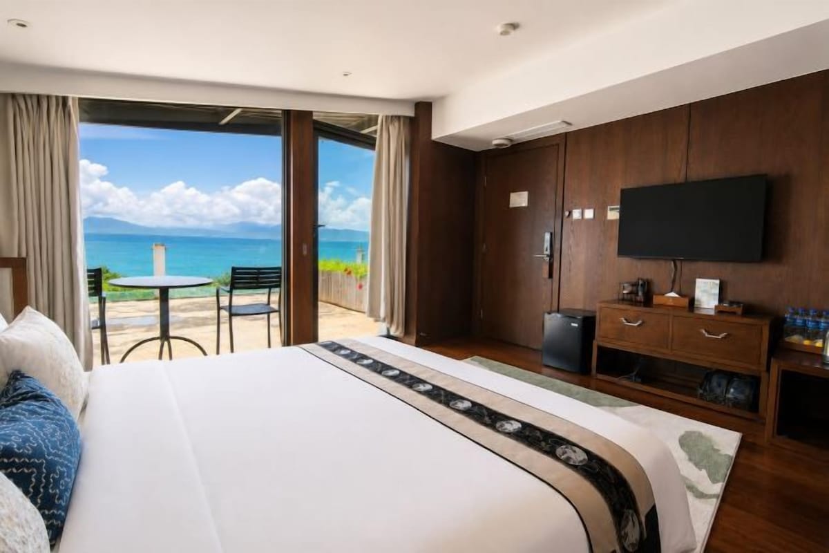 A hotel room with a bed, a television, a desk, and a view of the ocean.
