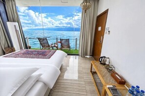A hotel room with a bed, a small table with a kettle, a chair, and a view of the sea.