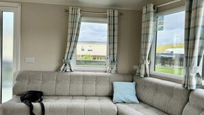 Un sofá seccional color beige, con un bolso negro y una almohada azul, dos ventanas con cortinas a cuadros, y vista a una casa y una valla.