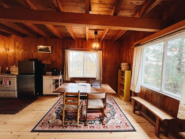 A wooden cabin interior with a dining table, chairs, a bench, a kitchen area with appliances, and large windows. - Classic Chelsea Cottage on the Gatineau River and along the famous Chelsea Trail (Chelsea)