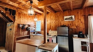 A rustic cabin interior with a wooden interior, a kitchen area including a stove, refrigerator, and bar stools.