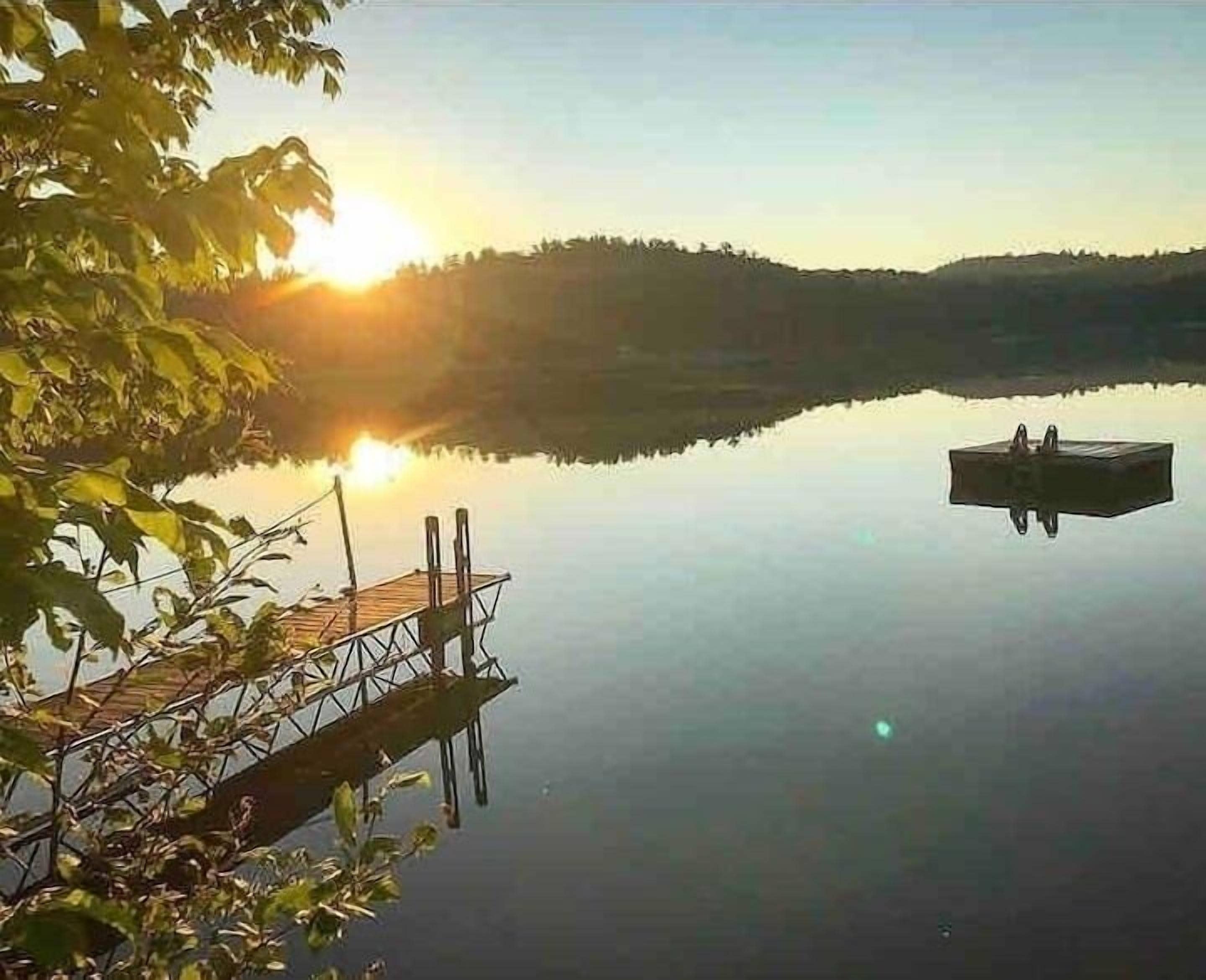 Ein ruhiger See mit einer hölzernen Anlegestelle, die ins Wasser ragt, umgeben von Bäumen und einer entfernten Hügelkette.