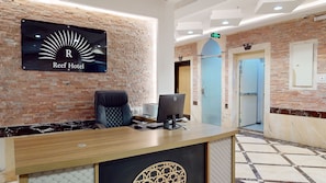 Un comptoir d’accueil d’hôtel avec un ordinateur, une chaise et une enseigne indiquant « Reef Hotel ». On y voit des portes et un sol carrelé.