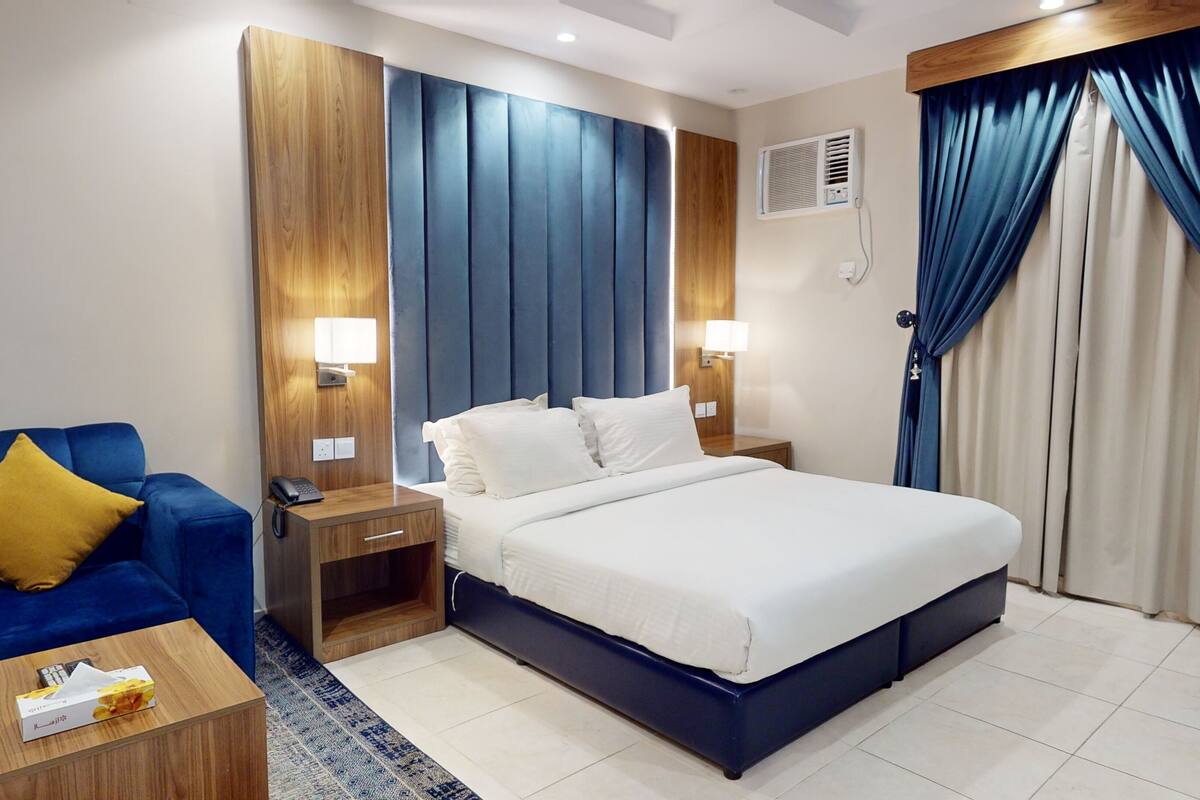 Une chambre d’hôtel avec un lit, un fauteuil bleu, une table de chevet et une commode.