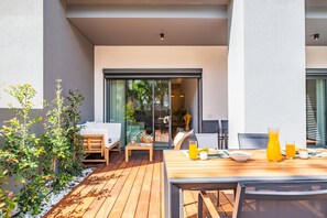 Terraza de madera con mesa preparada para el desayuno, un sofá y un ventanal que da a un espacio interior.