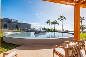 Ein moderner Poolbereich mit Holzterrasse und Liegestühlen, umgeben von Palmen und blauem Himmel.