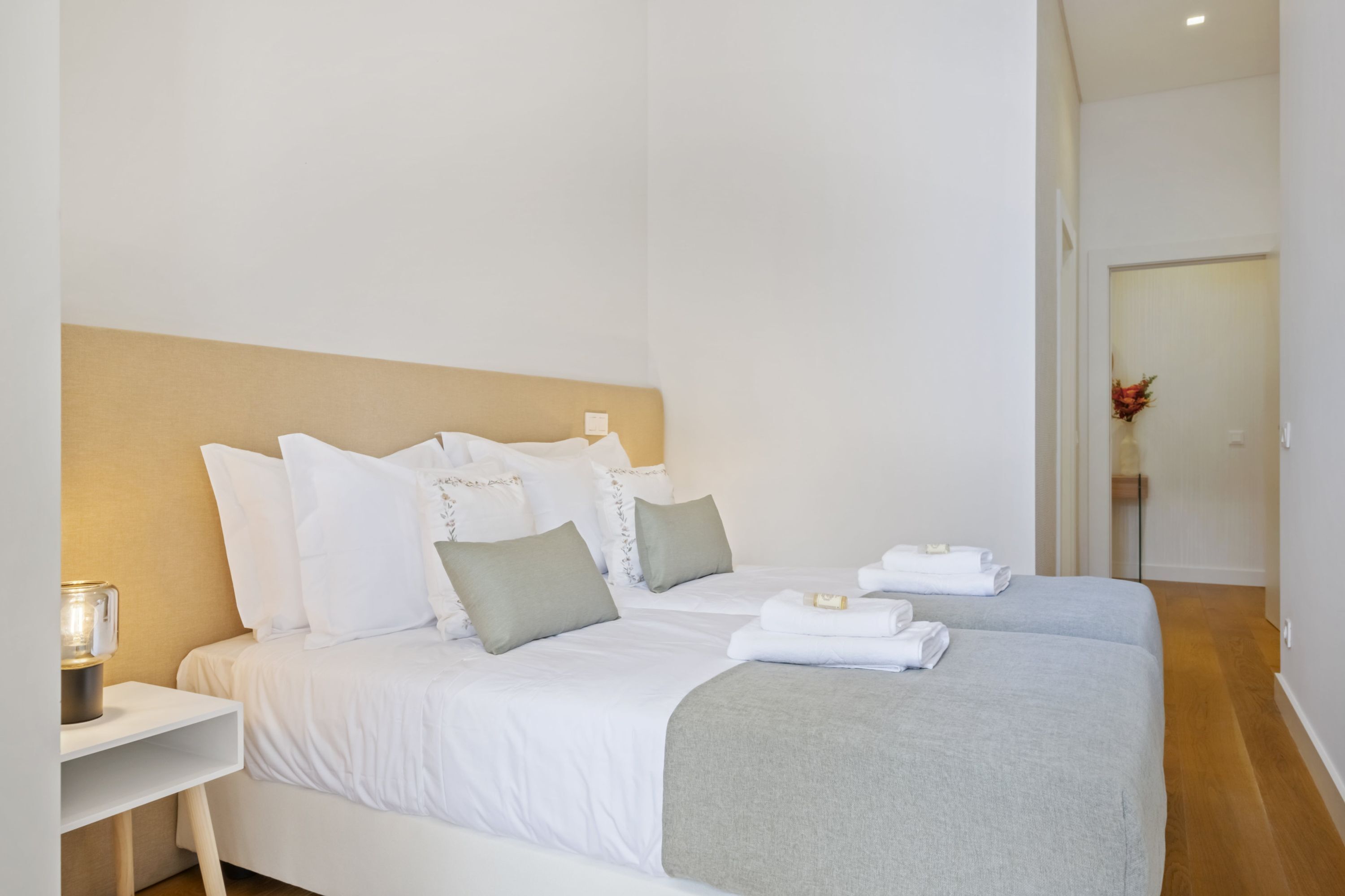 A hotel room with two beds, white bedding, a bedside table with a lamp, and a wooden floor.
