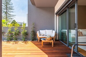 A modern outdoor patio with a wooden deck, a white sofa, and a glass door leading to a room.