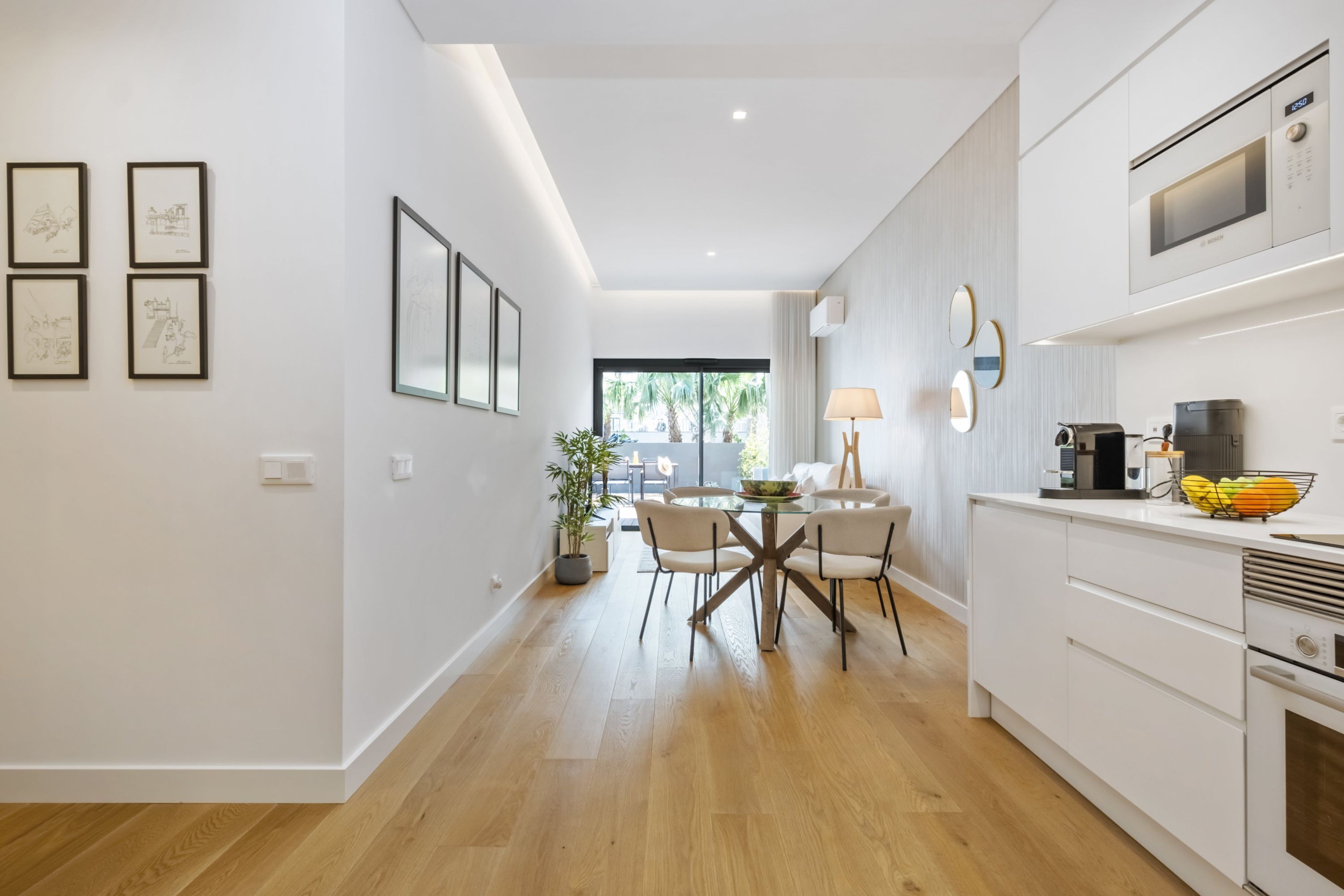 A modern kitchen with white cabinets, a microwave, and a coffee machine. There is a dining area with a table and chairs, and a large window providing natural light.