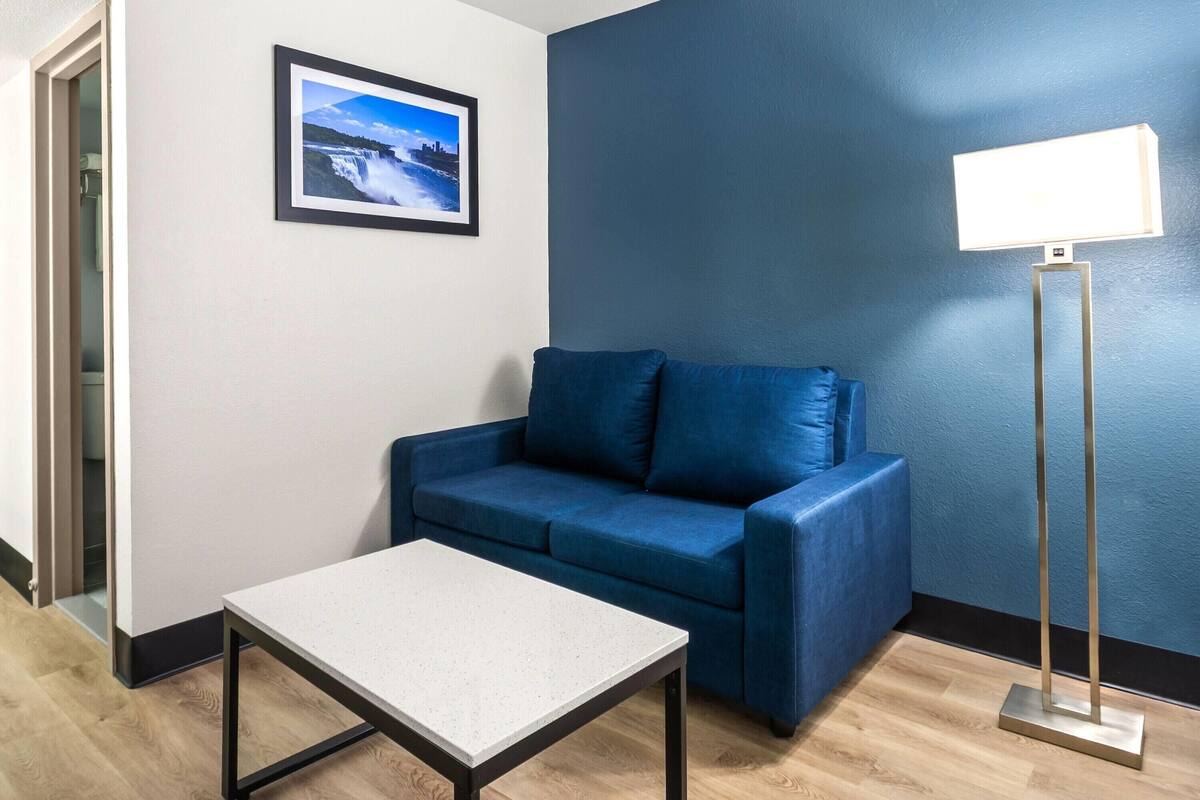 Una habitación con un sofá azul, una mesita pequeña, un cuadro enmarcado en la pared y una lámpara de pie.